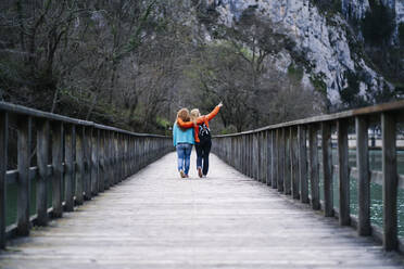 Back view of two best friends walking side by side on boardwalk, Valdemurio Reservoir, Asturias, Spain - DGOF00665