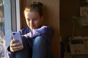 Porträt eines lächelnden Mädchens, das abends barfuß auf der Fensterbank sitzt und in sein Smartphone schaut - AUF00331