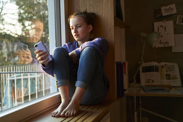 Girl sitting barefoot on window sill in the evening looking at smartphone - AUF00332