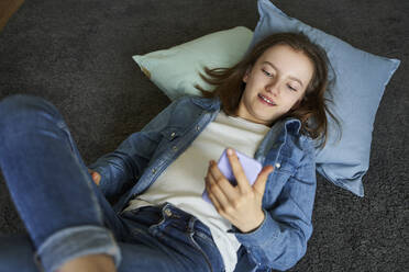Porträt eines Mädchens, das zu Hause auf dem Teppich liegt und auf sein Smartphone schaut - AUF00336