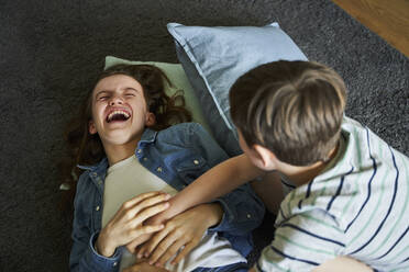 Junge kitzelt seine ältere Schwester, die zu Hause auf dem Teppich liegt - AUF00339