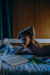 Hemdloser Junge, der ein Buch liest, während er zu Hause auf dem Bett liegt - EYF03470