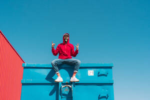 Young man wearing red hoodie, sitting on edge of blue container, making sign of the horns - ERRF03119