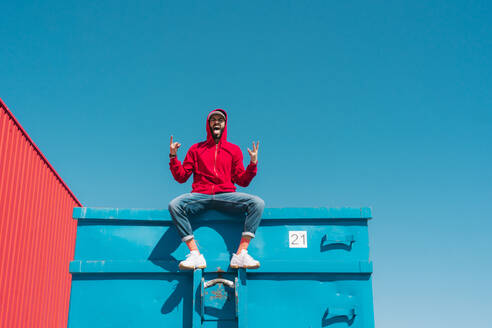 Junger Mann mit rotem Kapuzenpulli, der auf dem Rand eines blauen Containers sitzt und das Zeichen der Hörner macht - ERRF03119