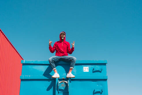 Young man wearing red hoodie, sitting on edge of blue container, making sign of the horns - ERRF03119