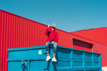 Young man wearing red hoodie, sitting on edge of blue container in front of red wall, raising forefinger - ERRF03127