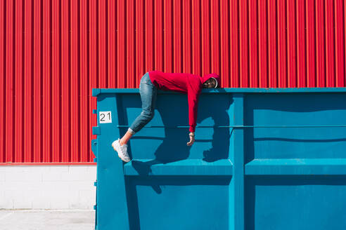 Junger Mann mit roter Kapuzenjacke liegt auf dem Rand eines Containers vor einer roten Wand - ERRF03129