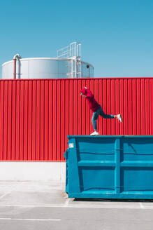 Young man wearing red hooded jacket balancing on edge of container in front of red wall in industrial setting - ERRF03131