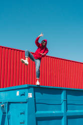 Young man wearing red hooded jacket balancing on edge of container in front of red wall - ERRF03132