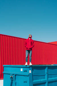 Junger Mann in roter Kapuzenjacke steht auf dem Rand eines Containers vor einer roten Wand - ERRF03133