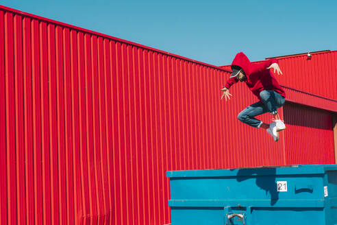 Young man wearing red hooded jacket jumping from edge of container in front of red wall - ERRF03135