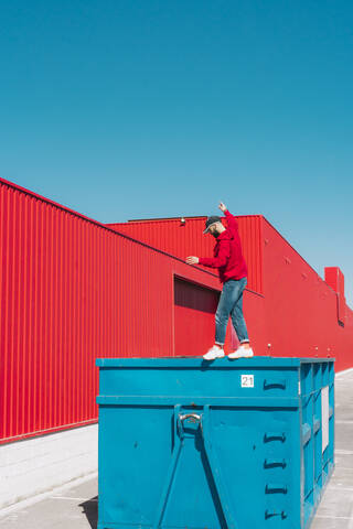 Junger Mann in roter Kapuzenjacke balanciert auf dem Rand eines Containers vor einer roten Wand, lizenzfreies Stockfoto