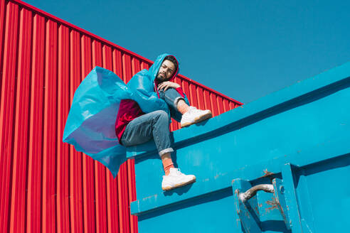 Junger Mann in Regenjacke sitzt auf dem Rand eines blauen Containers vor einer roten Wand - ERRF03141