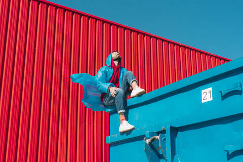 Junger Mann in Regenjacke sitzt auf dem Rand eines blauen Containers vor einer roten Wand, lizenzfreies Stockfoto