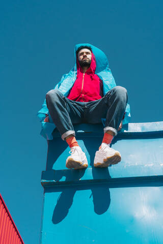 Junger Mann in roter Kapuzenjacke sitzt auf einem Container vor blauem Himmel, lizenzfreies Stockfoto