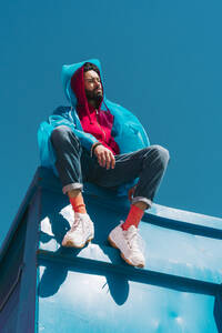 Junger Mann sitzt auf dem Rand eines blauen Containers und trägt einen Regenmantel - ERRF03145