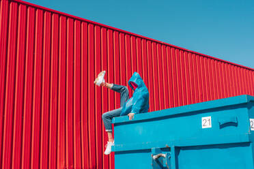 Junger Mann in Regenjacke sitzt auf dem Rand eines blauen Containers vor einer roten Wand und hebt das Bein - ERRF03146