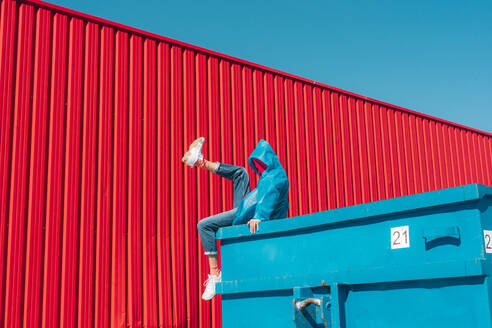 Junger Mann in Regenjacke sitzt auf dem Rand eines blauen Containers vor einer roten Wand und hebt das Bein - ERRF03146