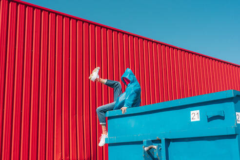 Young man wearing rain coat sitting on edge of blue container in front of red wall, raising leg - ERRF03146