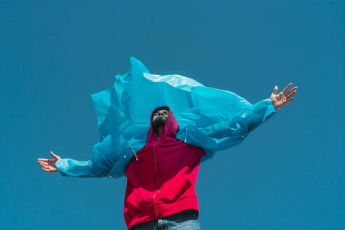 Young man wearing plastic rain coat, stanind in gthe wind with arms outstretched - ERRF03147