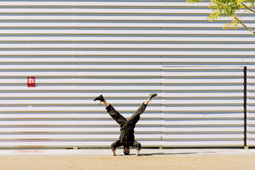 Mann in schwarzem Overall macht Handstand vor einem Industriegebäude - ERRF03154