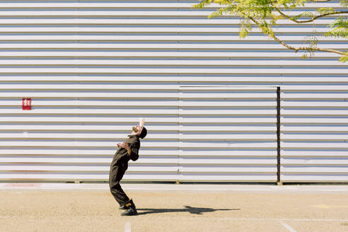 Man wearing black overall balancing cup on his forehead in front of industrial building - ERRF03164