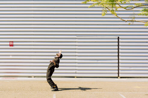 Mann in schwarzem Overall balanciert Tasse auf der Stirn vor einem Industriegebäude - ERRF03164