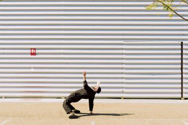 Mann in schwarzem Overall balanciert Tasse auf der Stirn vor einem Industriegebäude - ERRF03165