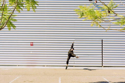 Man wearing black overall jumping in the air in front of industrial building - ERRF03170