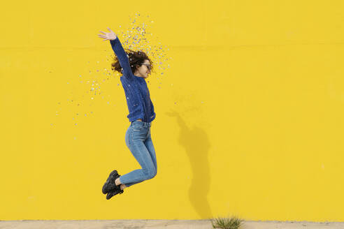 Young woman jumping in the air in front of yellow background throwing confetti - JCZF00034