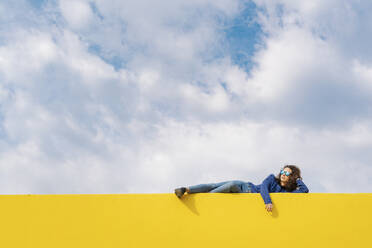 Young woman relaxing on yellow wall against cloudy sky - JCZF00035