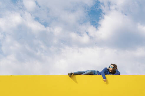 Junge Frau entspannt sich auf gelber Wand gegen bewölkten Himmel - JCZF00035