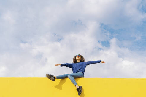 Glückliche junge Frau sitzt auf gelber Wand gegen bewölkten Himmel und schaut nach oben - JCZF00036
