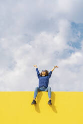 Glückliche junge Frau sitzt auf gelber Wand gegen bewölkten Himmel und schaut nach oben - JCZF00037