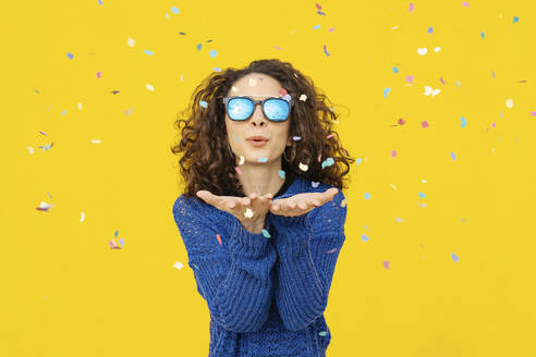 Portrait of young woman with mirrored sunglasses blowing confetti in the air in front of yellow background - JCZF00041