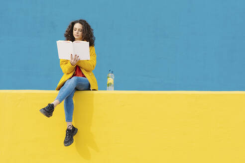 Young woman sitting on yellow wall reading a book - JCZF00046