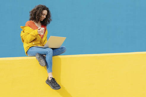 Happy young woman with coffee to go sitting on yellow wall using laptop - JCZF00047