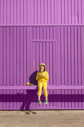 Kleines, gelb gekleidetes Mädchen sitzt auf einer Bar vor einem lila Garagentor und trinkt - ERRF03171