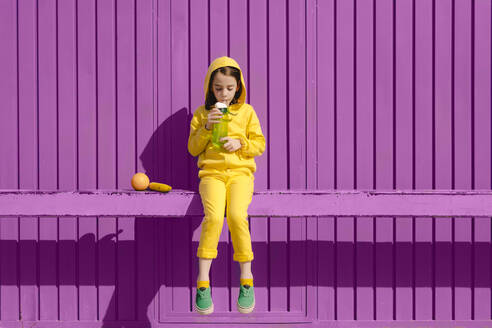 Kleines Mädchen in gelb gekleidet sitzt auf Bar vor lila Hintergrund trinken - ERRF03172