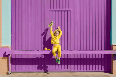 Glückliches kleines Mädchen in gelber Kleidung, das auf einer Stange vor einem lila Garagentor sitzt - ERRF03173