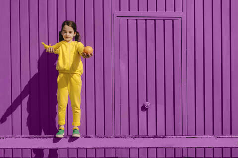 Porträt eines kleinen, gelb gekleideten Mädchens, das vor einem lila Hintergrund steht und Früchte anbietet, lizenzfreies Stockfoto