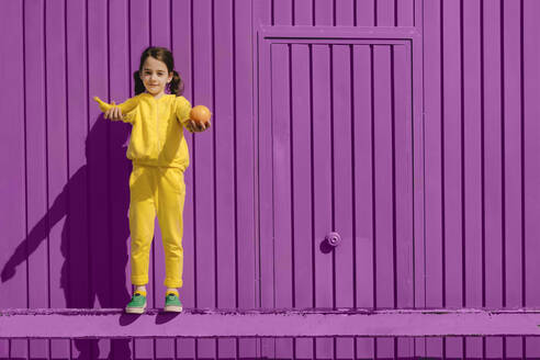 Porträt eines kleinen, gelb gekleideten Mädchens, das vor einem lila Hintergrund steht und Früchte anbietet - ERRF03176