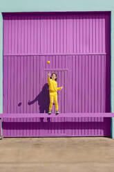 Happy little girl dressed in yellow balancing on bar in front of purple garage throwing orange in the air - ERRF03180