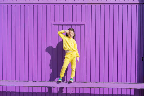 Kleines, gelb gekleidetes Mädchen steht auf einer Stange vor einem lila Garagentor - ERRF03181