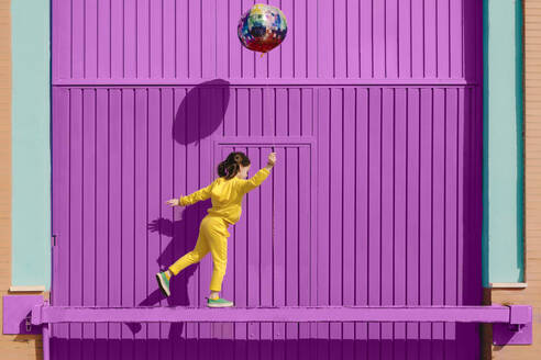 Little girl dressed in yellow balancing on bar in front of purple garage door holding balloon - ERRF03182