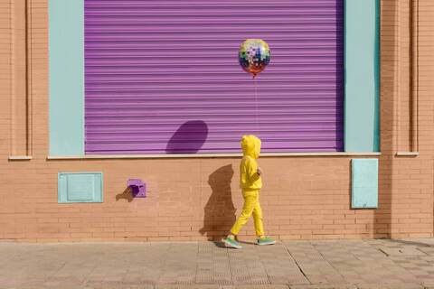 Kleines, gelb gekleidetes Mädchen geht mit Luftballon auf dem Bürgersteig, lizenzfreies Stockfoto