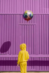 Rückenansicht von gelb gekleideten Kindern, die einen Luftballon vor einem lila Hintergrund halten - ERRF03185
