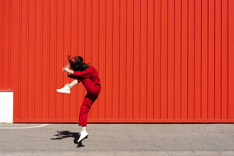 Woman dressed in red overall jumping in the air in front of red roller shutter stock photo