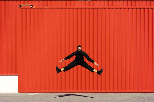 Lächelnder, schwarz gekleideter Mann, der vor einem roten Rolltor in die Luft springt - ERRF03191