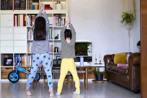 Rückenansicht von zwei Schwestern, die auf einen Laptop schauen, während sie im Wohnzimmer trainieren - ERRF03199
